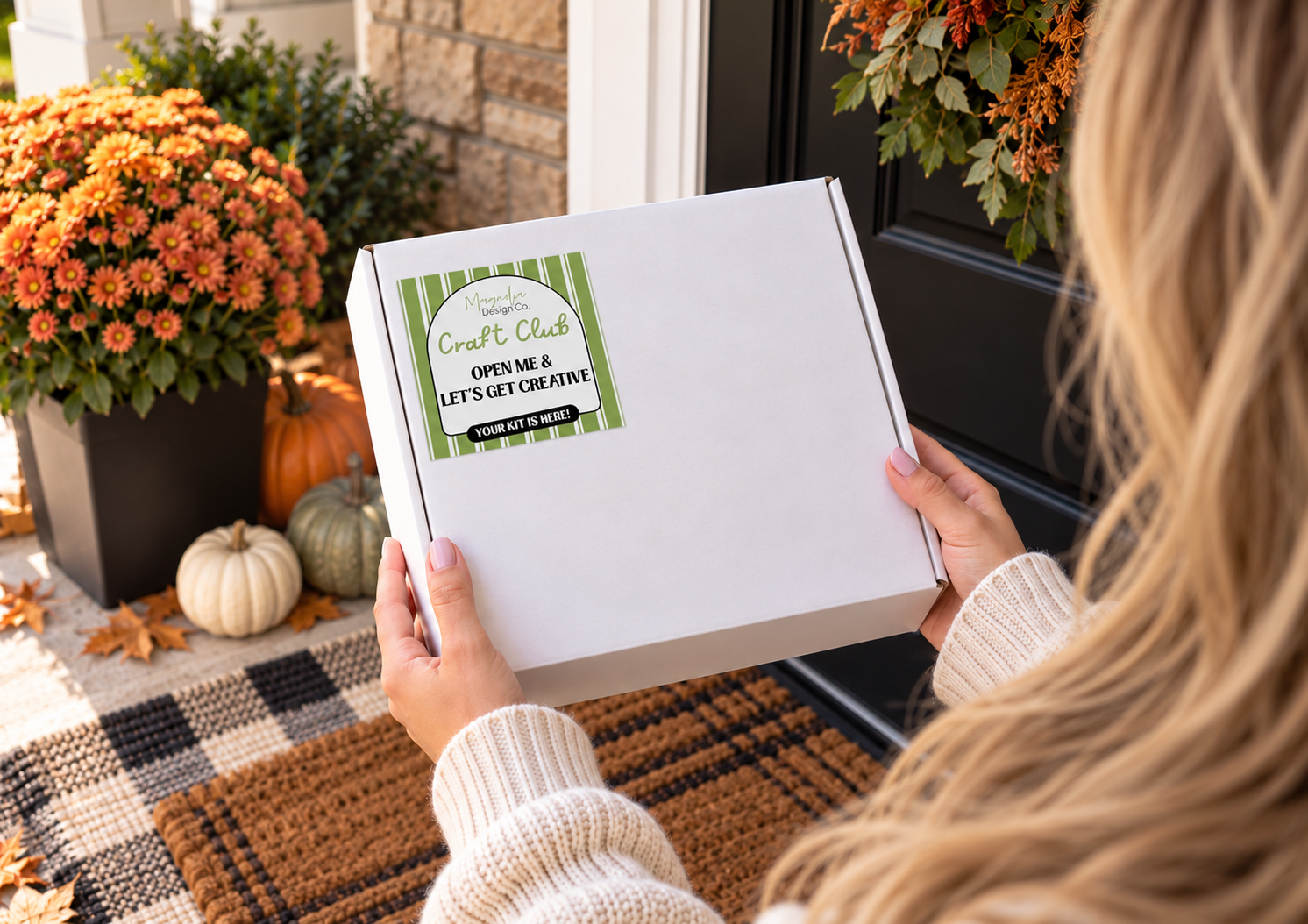 A Craft Club box held in two hands on a front porch, with orange mums, pumpkins and fallen leaves beside a plaid doormat and a black front door